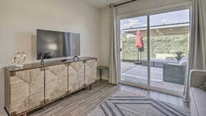 Een moderne woonkamer met een flatscreen televisie op een houten kast, en een schuifdeur naar een buitenpatio met meubilair.