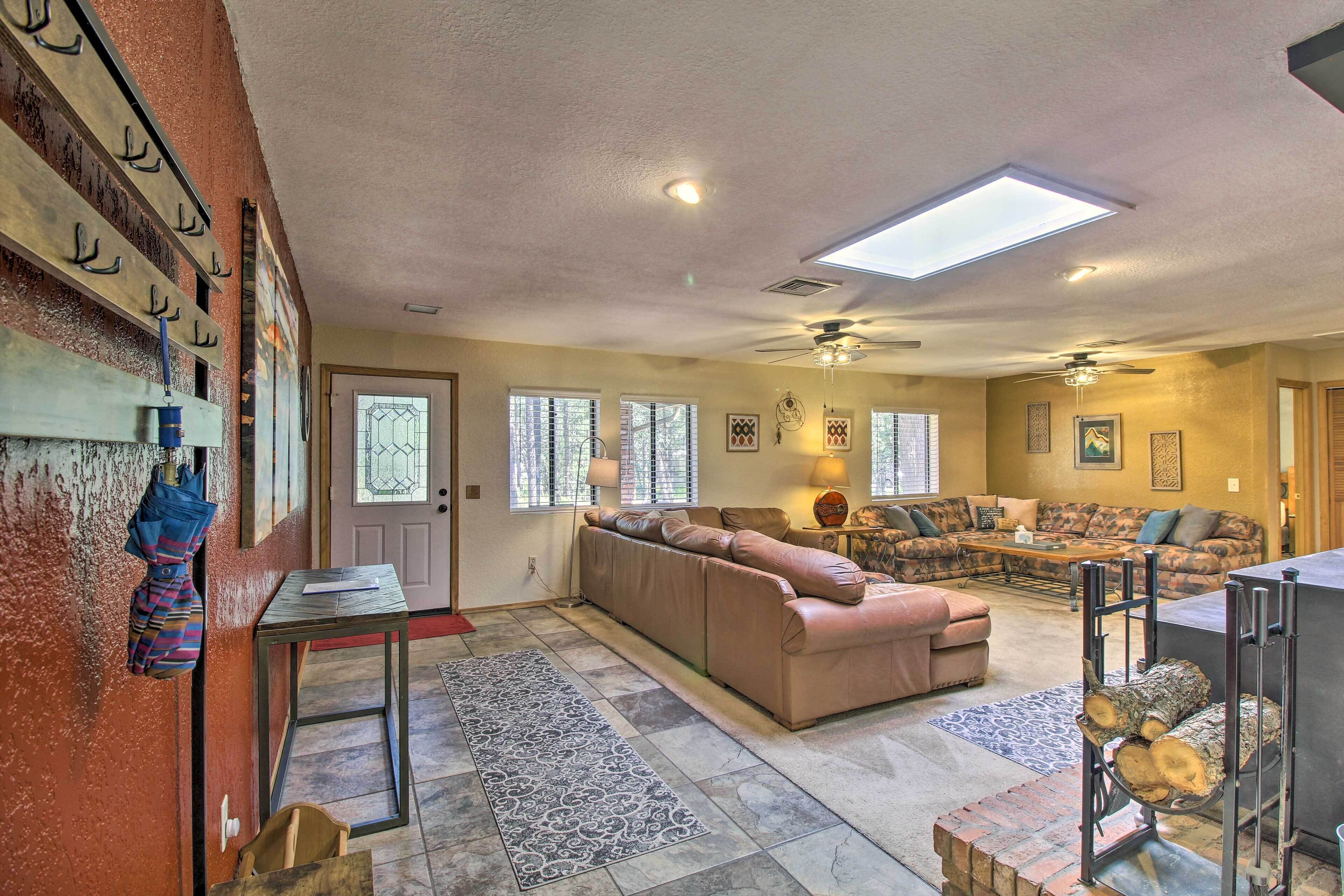 A spacious living room with a large sectional sofa, a coffee table, and a fireplace. There are multiple windows providing natural light and a ceiling fan.