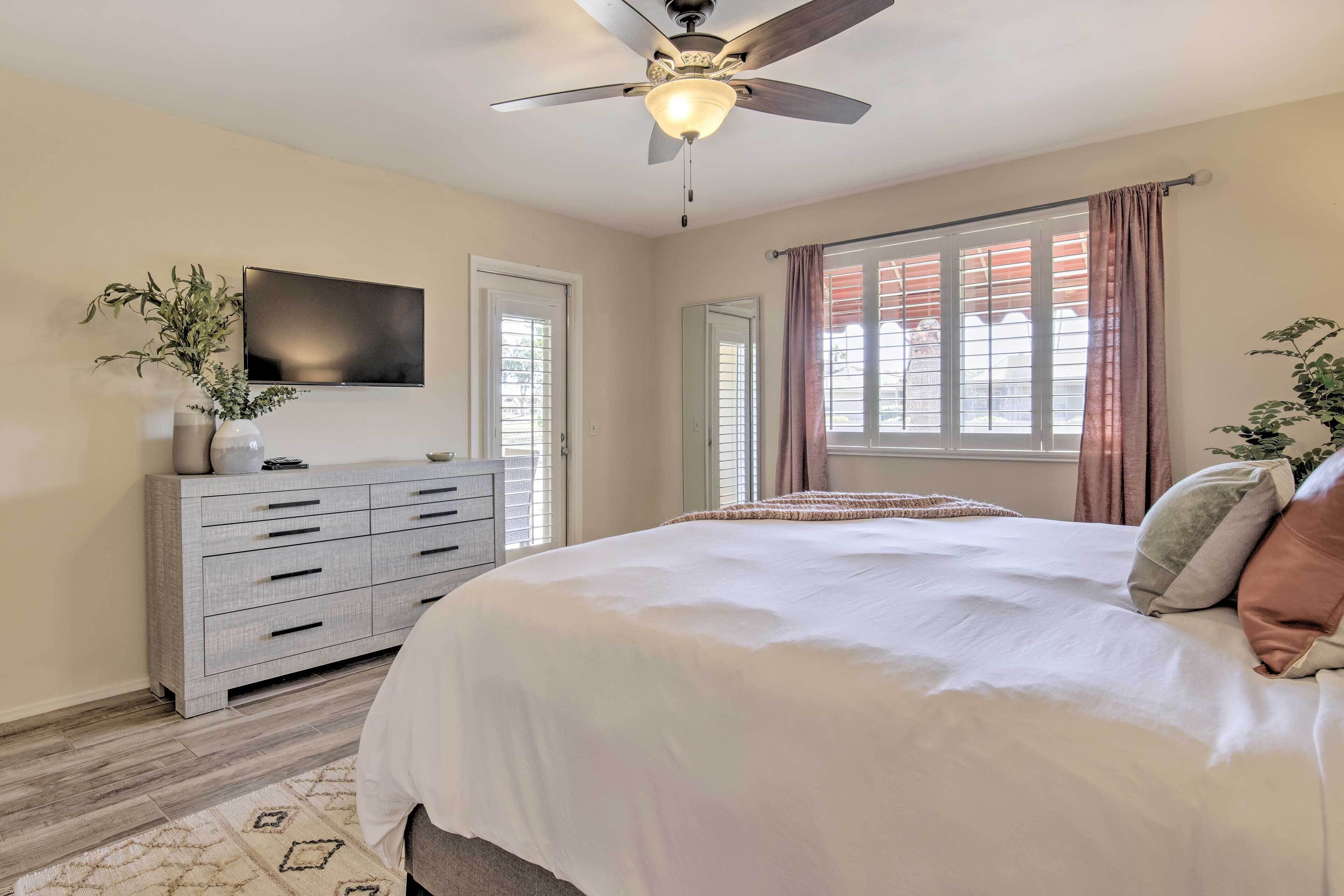 A bedroom with a bed, a ceiling fan, a TV mounted on the wall, a dresser, and a window with curtains.