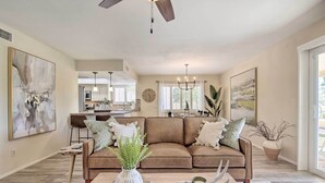 A modern living room with a brown leather sofa, a coffee table with books and a vase, and a dining area in the background. - Stunning Sun Lakes Home w/ Beautiful Lake Views! (Chandler)