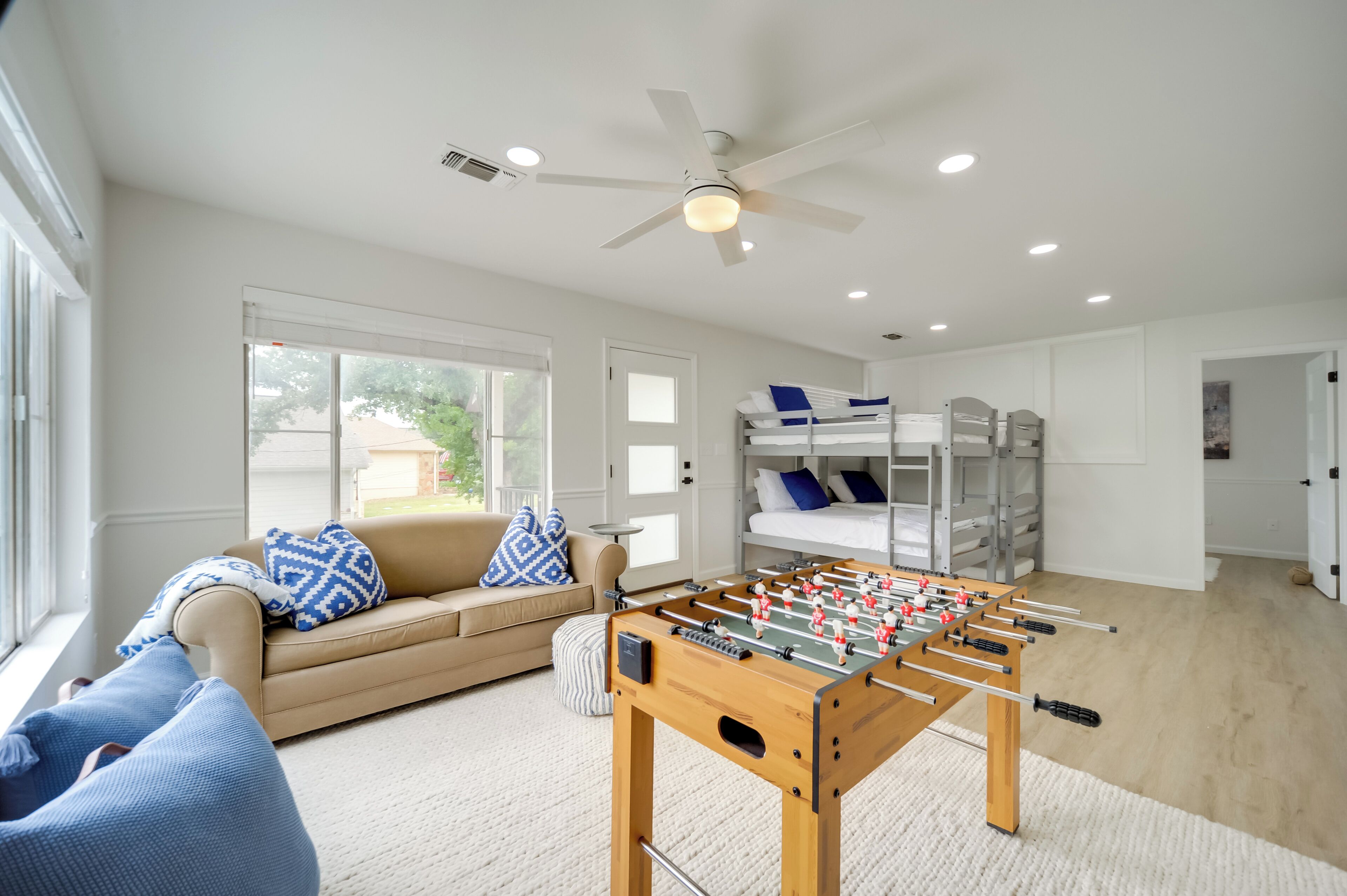 A modern living room with a foosball table, a sofa with patterned cushions, a ceiling fan, and a bunk bed with pillows.