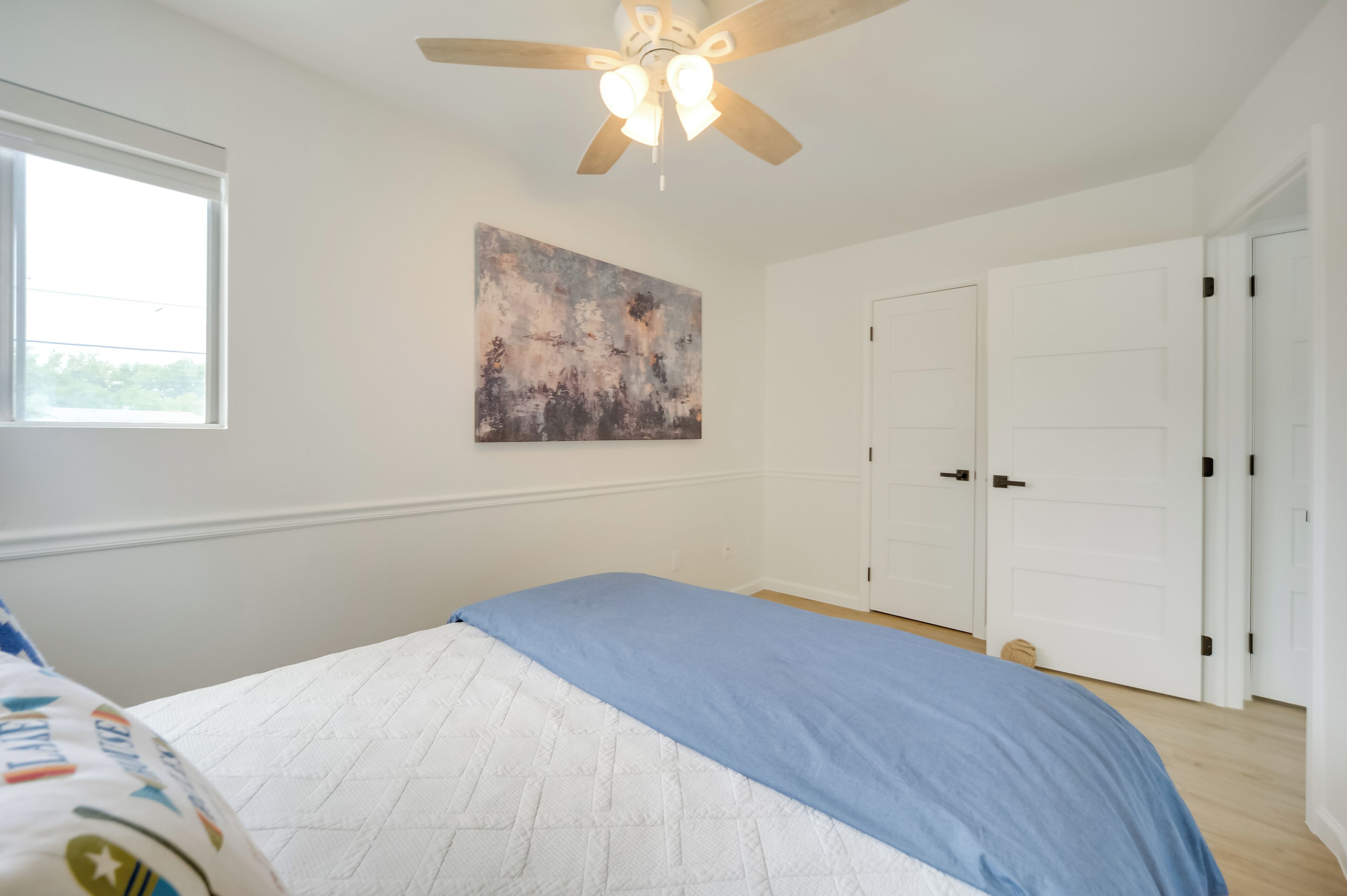 A bedroom with a bed, a ceiling fan, a window, a door, and a painting on the wall.