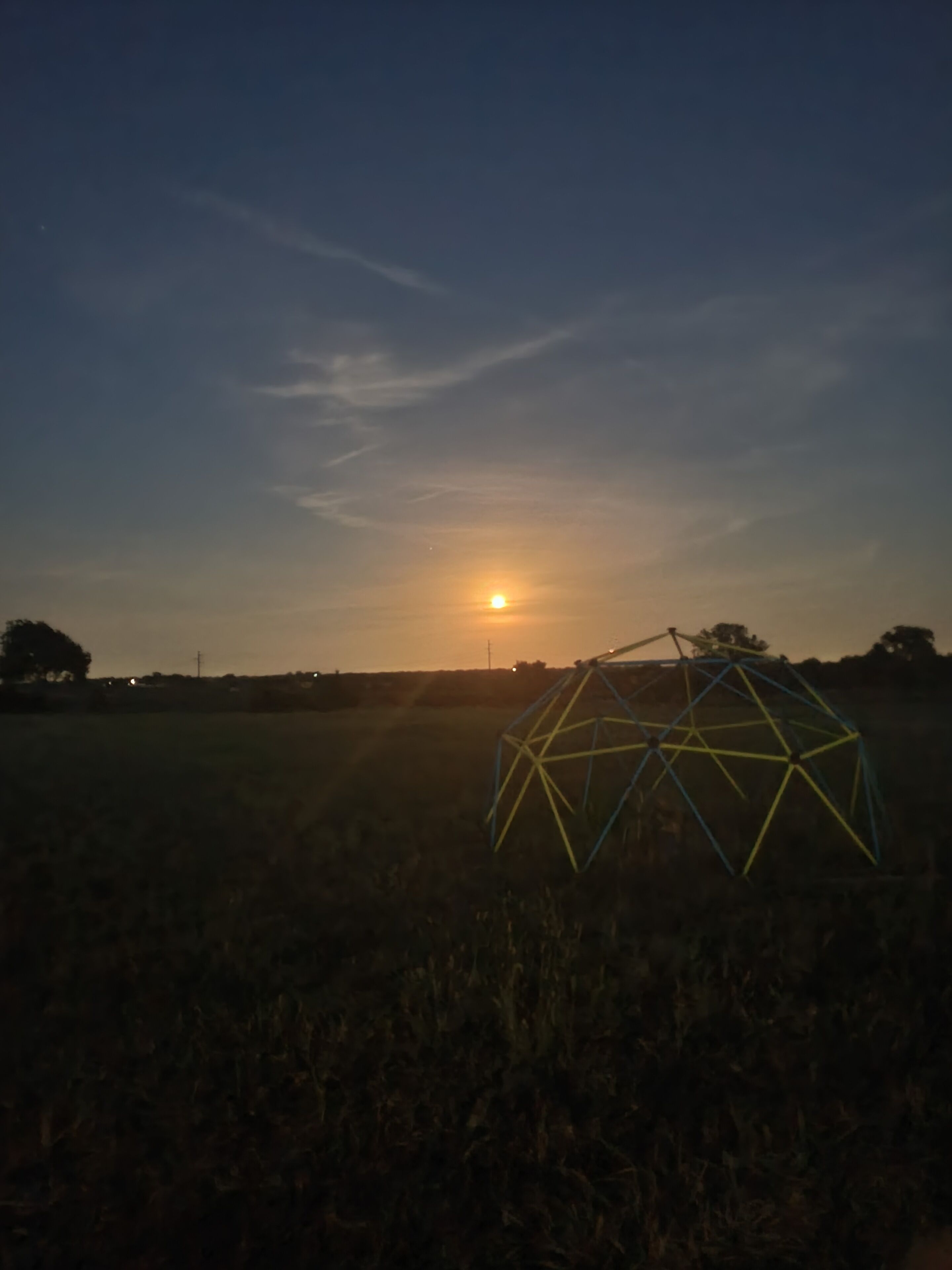 Une structure en forme de dôme géodésique se dresse dans un champ au coucher du soleil.