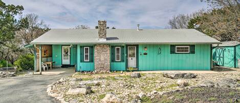 A single-story house with a stone chimney, green exterior, and a front porch with a table and chairs.