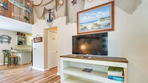 A living room with a wooden floor, a TV on a wooden stand, a bookshelf, and a painting of a beach scene. - Sun, Sand & Smiles! Ocean Isle Apt w/ Balcony (Ocean Isle Beach)