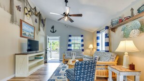 A living room with a TV, wooden floor, blue patterned chair, and nautical decor. - Sun, Sand & Smiles! Ocean Isle Apt w/ Balcony (Ocean Isle Beach)