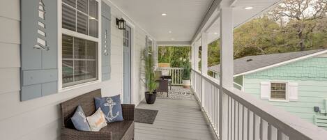 A covered porch with a wicker bench, potted plants, and a view of a green house.