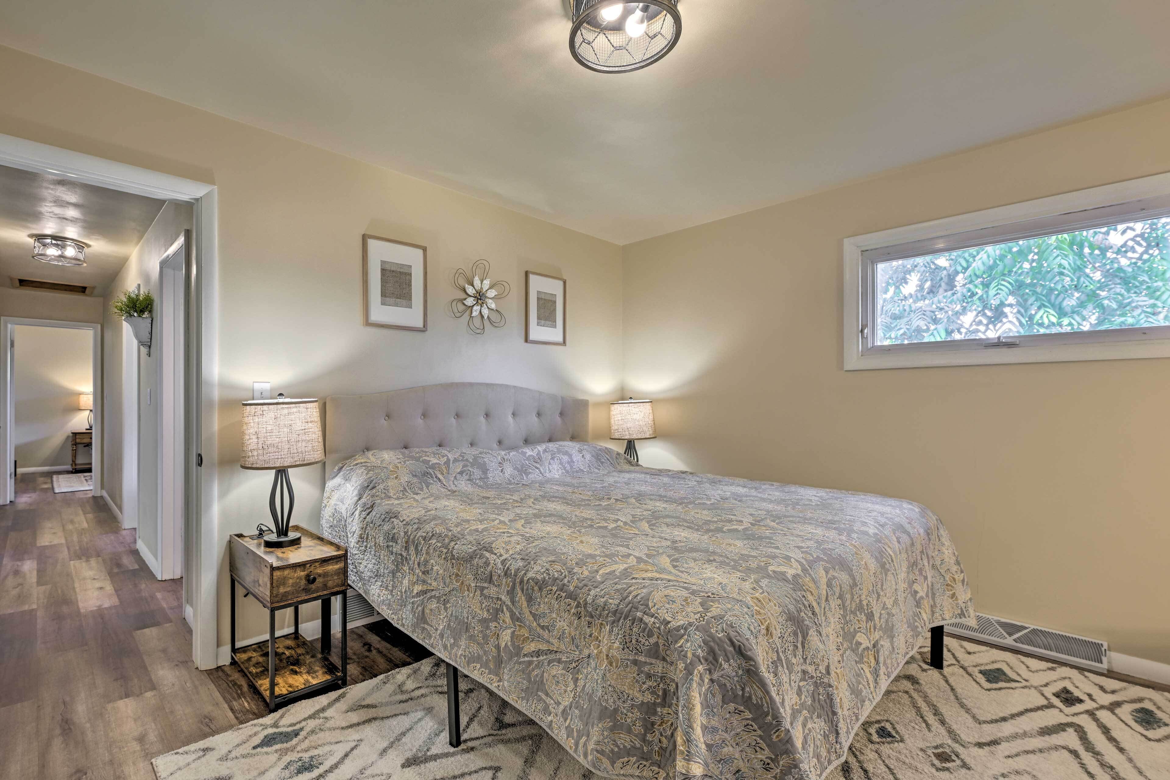 A bedroom with a large bed, two bedside lamps, a window with a view of trees, and a door leading to another room.