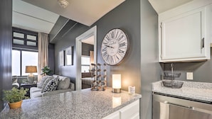 Una cocina moderna con encimera de granito, muebles blancos, un reloj de pared grande y una zona de comedor con sofá y lámpara.
