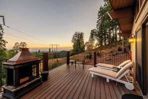 Una terraza de madera con chimenea, asientos exteriores y vistas a árboles y colinas.