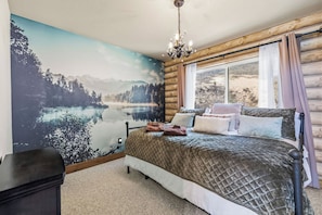 A bedroom with a large mural of a lake and mountains, a bed with multiple pillows, a chandelier, and a window with curtains. - Hot Tub + Scenic Views: Luxury Twain Harte Cabin! (Twain Harte)