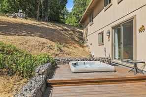A wooden deck with a hot tub, surrounded by a stone wall and greenery, leading to a house with a large sliding glass door. - Hot Tub + Scenic Views: Luxury Twain Harte Cabin! (Twain Harte)