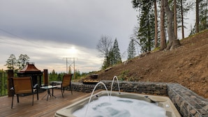 A hot tub on a wooden deck with chairs and a barbecue area, surrounded by a stone wall and trees. - Hot Tub + Scenic Views: Luxury Twain Harte Cabin! (Twain Harte)