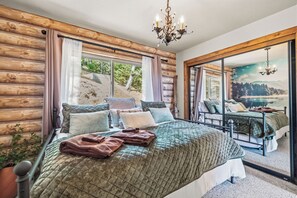 A bedroom with a large bed, multiple pillows, a view of a lake through a sliding door, and a chandelier. - Hot Tub + Scenic Views: Luxury Twain Harte Cabin! (Twain Harte)