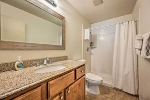 Une salle de bain avec un lavabo surmonté d’un plan de travail en granit, des meubles en bois, des toilettes blanches et une douche avec un rideau blanc.