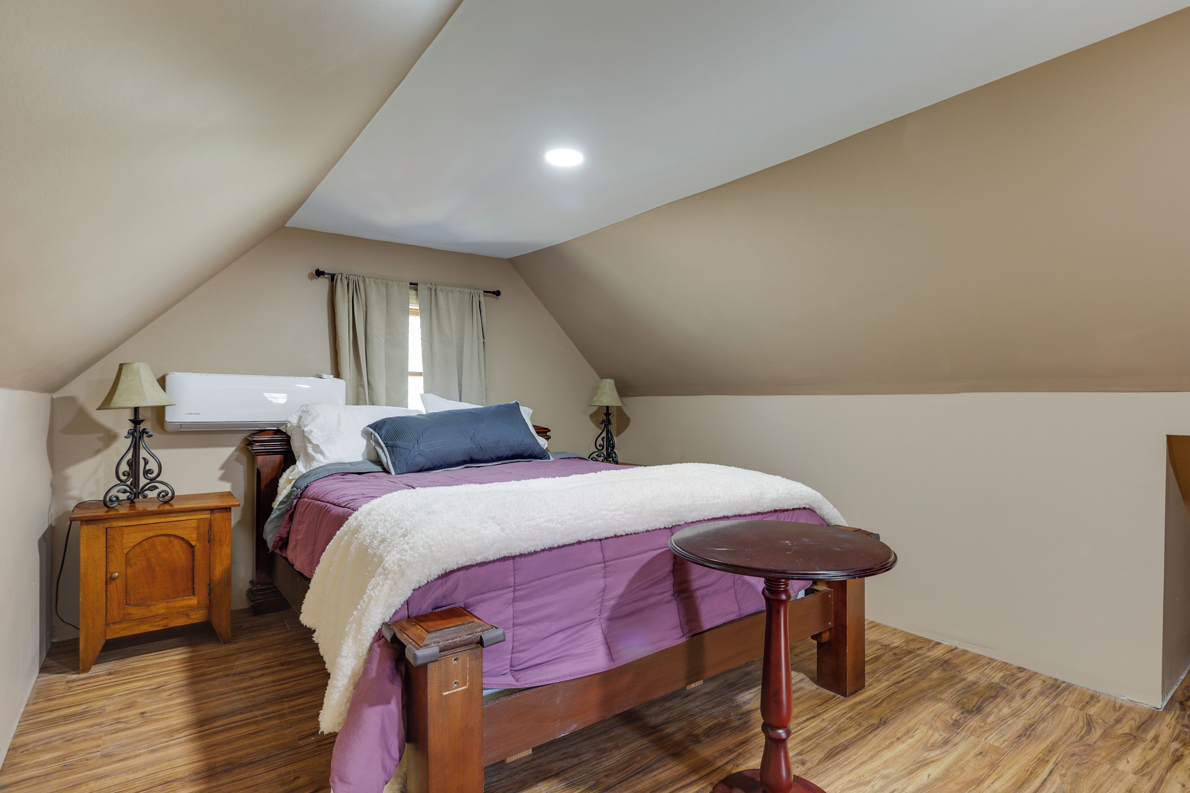 Un dormitorio con techo inclinado, una cama con colcha morada, una mesita de noche de madera, una pequeña mesa y un calefactor fijado a la pared.