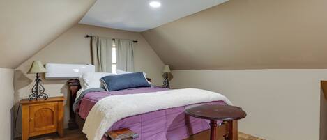 Un dormitorio con techo inclinado, una cama con colcha morada, una mesita de noche de madera, una pequeña mesa y un calefactor fijado a la pared.
