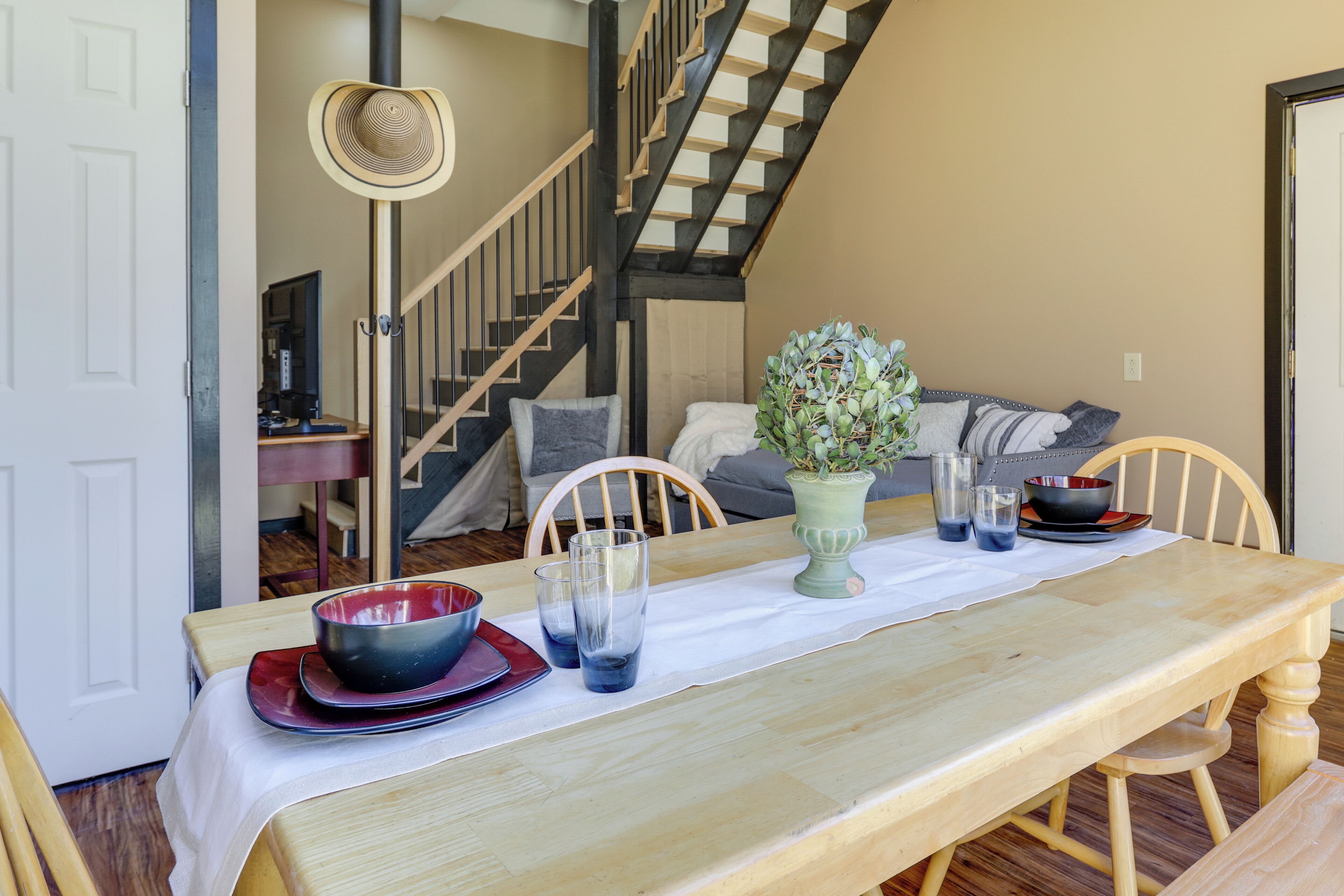 A dining area with a wooden table set for two, a staircase, and a living room with a sofa and cushions.