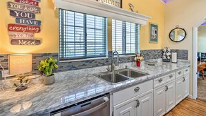 A kitchen with a marble countertop, stainless steel sink, and white cabinets. There is a window with blinds, a potted plant, and a wall-mounted mirror.