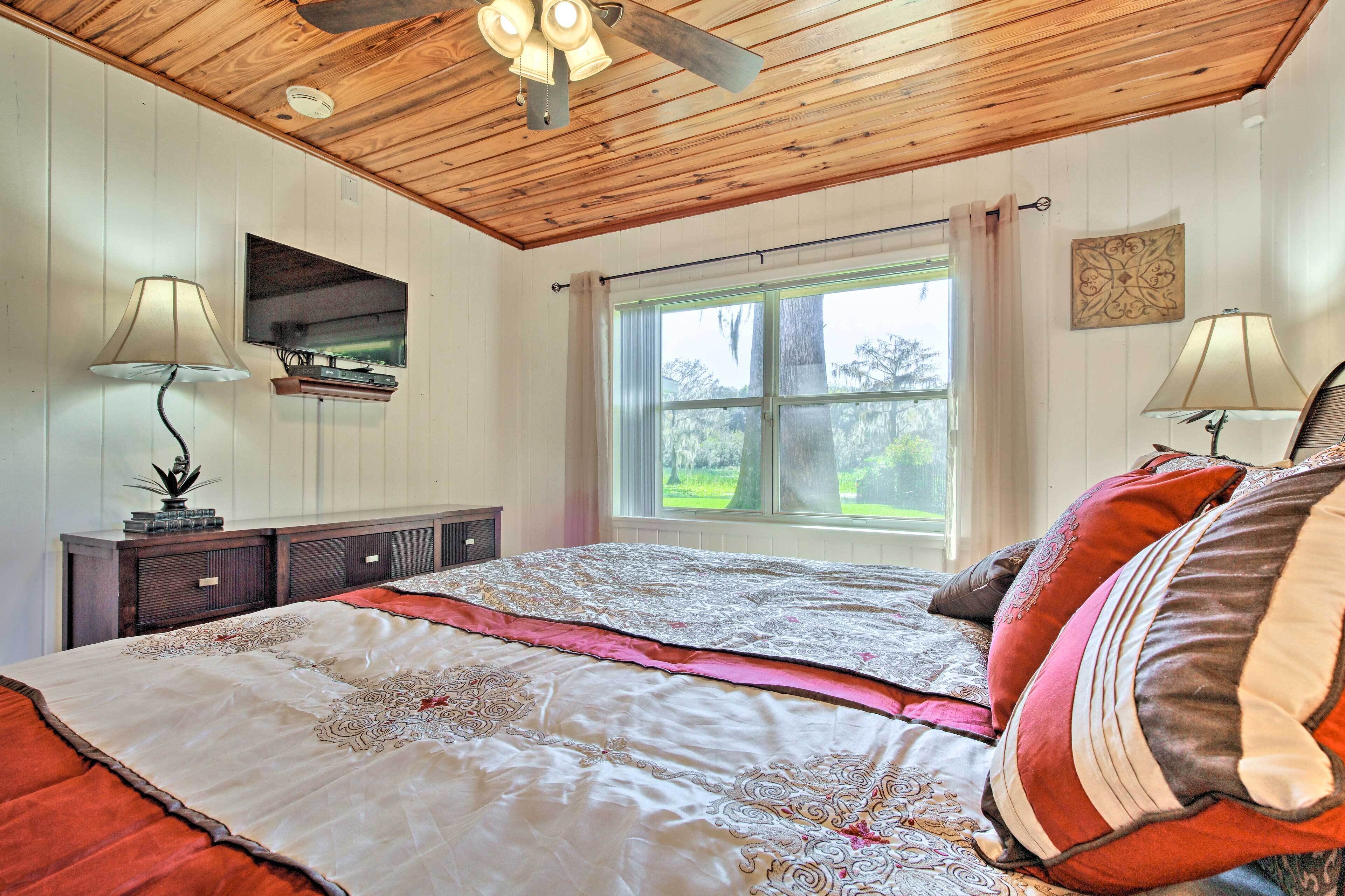 A bedroom with a wooden ceiling, a large bed with patterned bedding, a TV mounted on the wall, and a window with curtains.