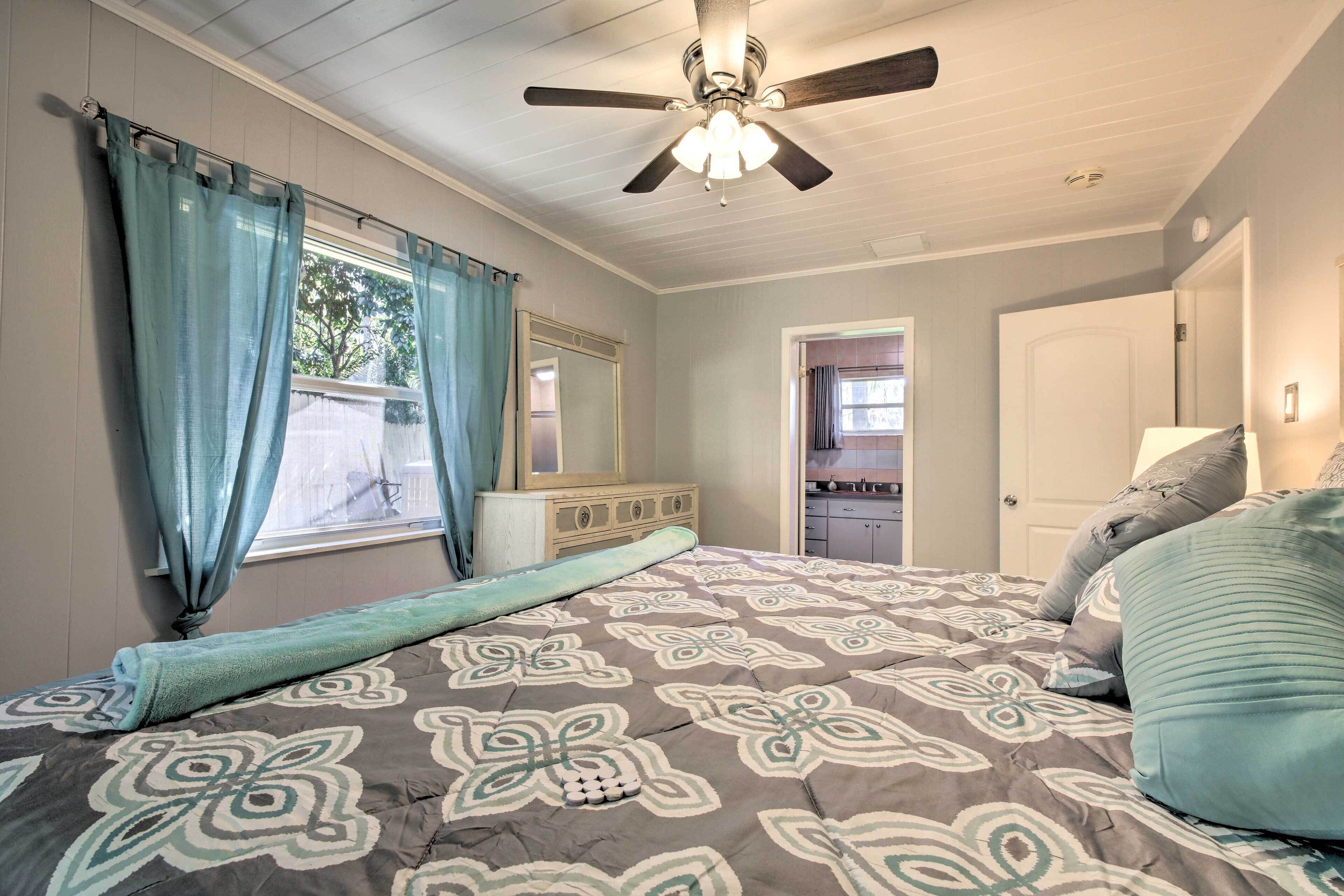 A bedroom with a bed, a ceiling fan, a window with curtains, a dresser, and a door leading to another room.
