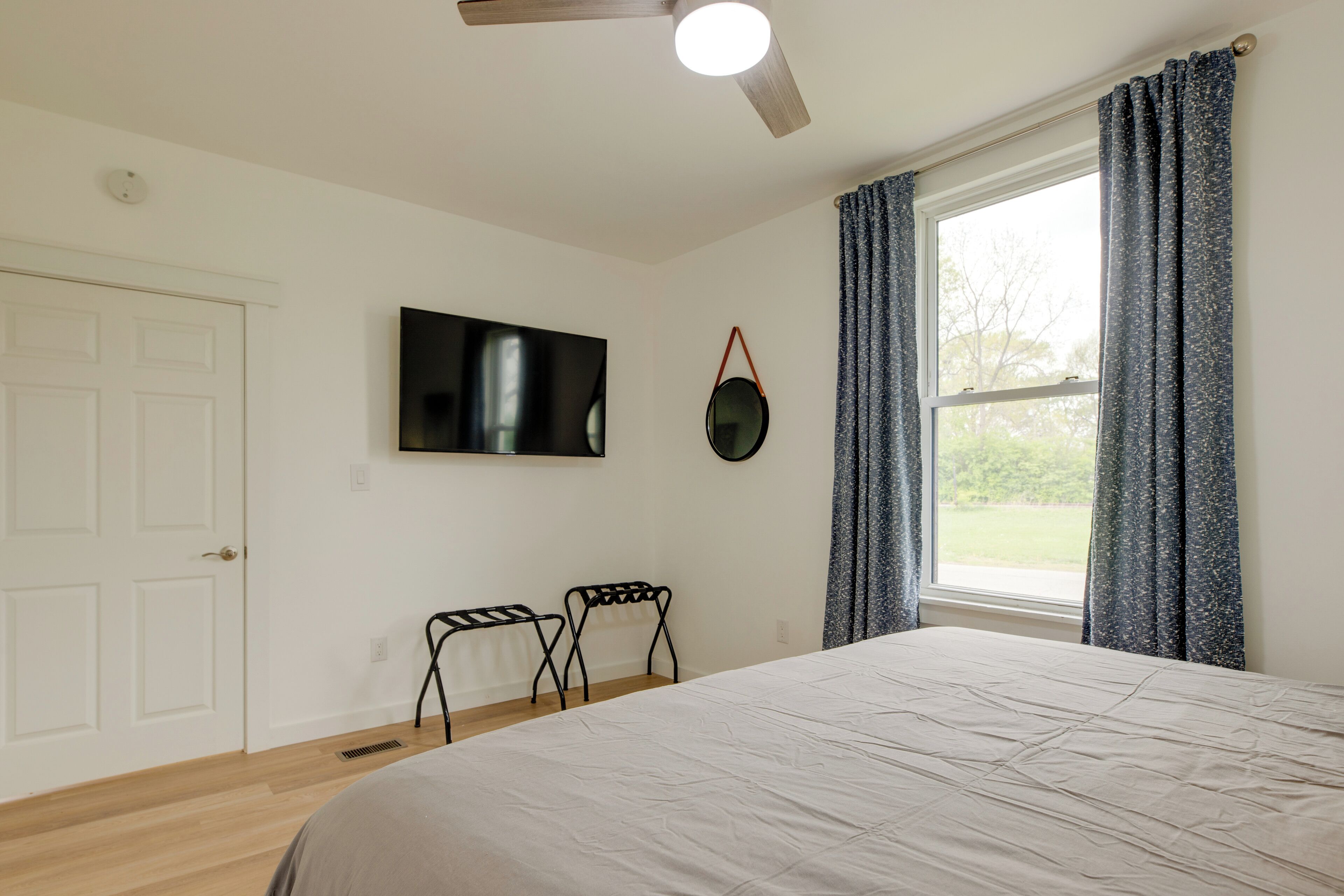 Ein Schlafzimmer mit Bett, Deckenventilator, an der Wand montiertem Fernseher und einem Fenster mit Vorhängen.