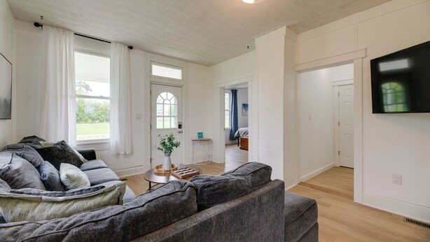 A spacious living room with a grey sofa, a coffee table, and a TV mounted on the wall. There is a door leading to another room and a window with curtains. - Modern Family Home in Mason w/ Backyard + Fire Pit (Mason)