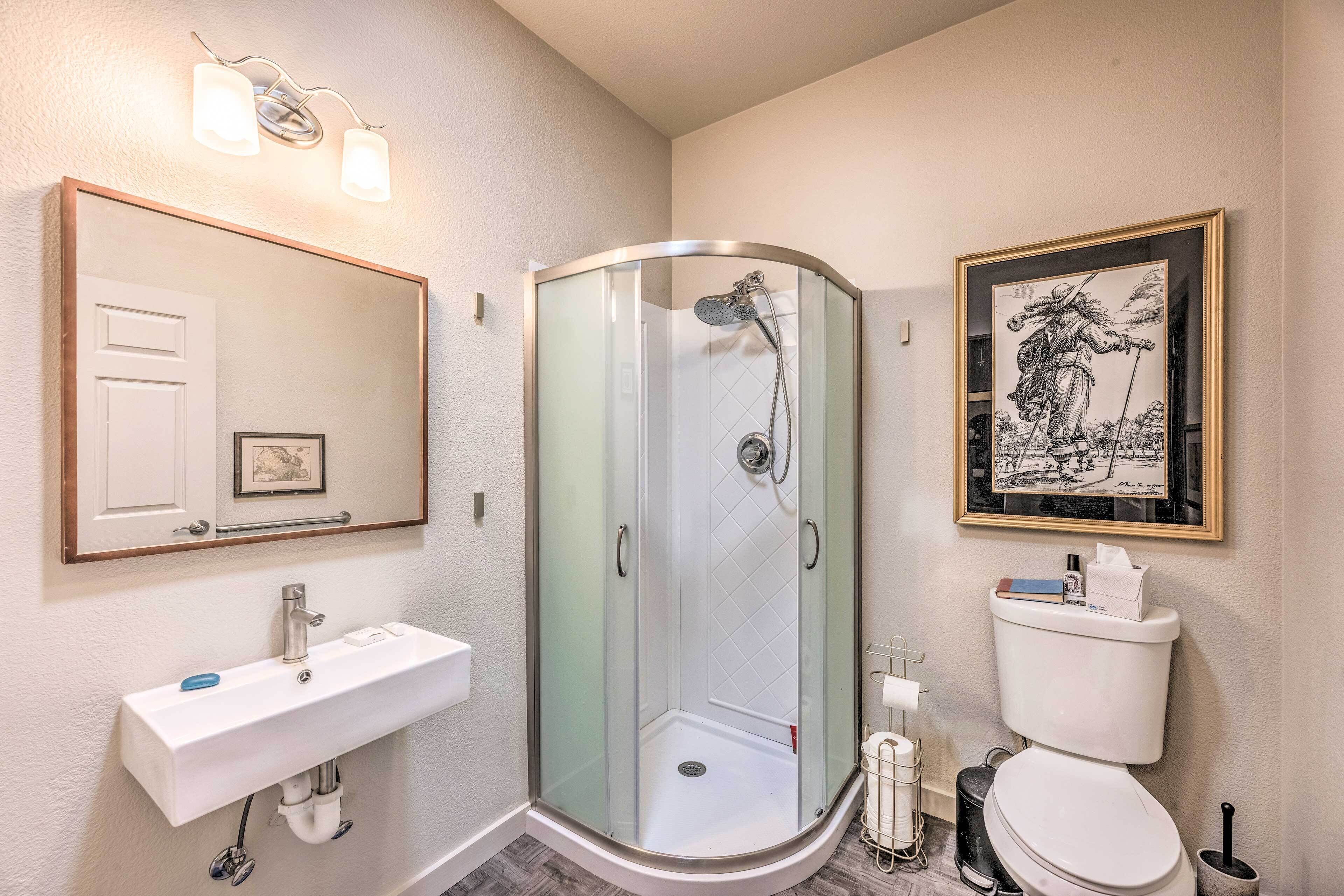 A bathroom with a glass-enclosed shower, a white sink with a mirror above it, and a toilet with a framed picture above it.