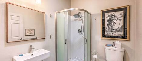A bathroom with a glass-enclosed shower, a white sink with a mirror above it, and a toilet with a framed picture above it.