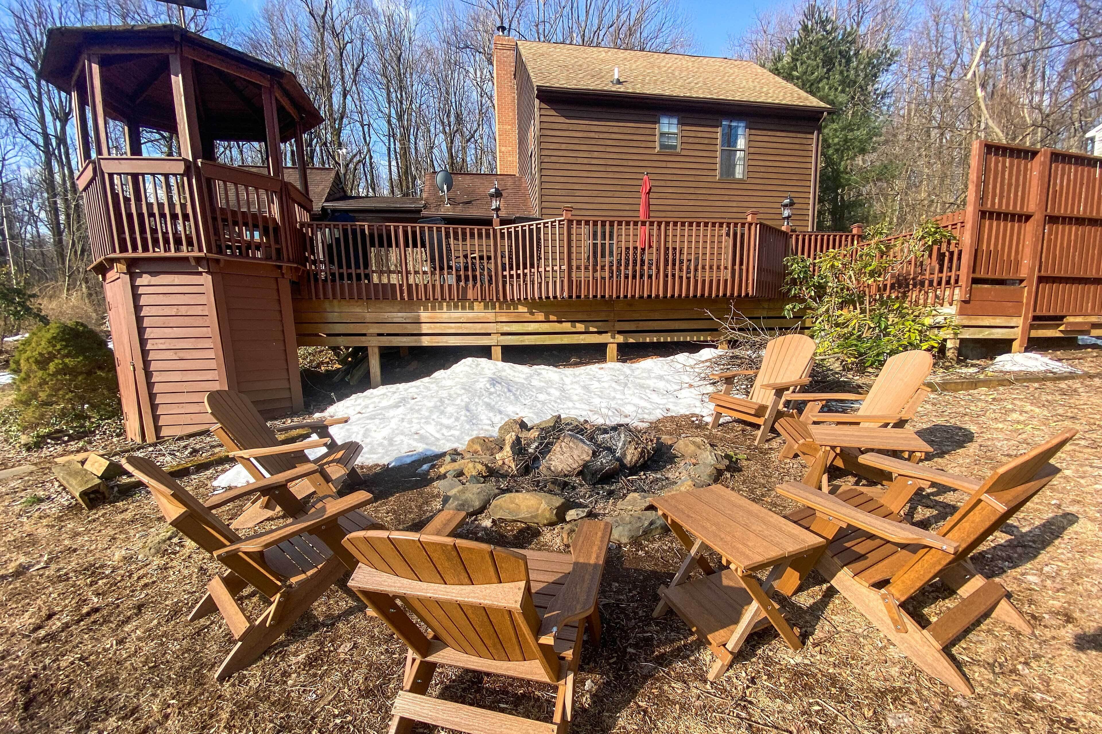 En træterrasse med Adirondack-stole og en bålplads, omgivet af terrasse og hus.