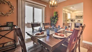 Un comedor con una mesa de madera dispuesta para cuatro personas, rodeada de sillas. Una araña cuelga encima, y un ventanal grande proporciona luz natural.