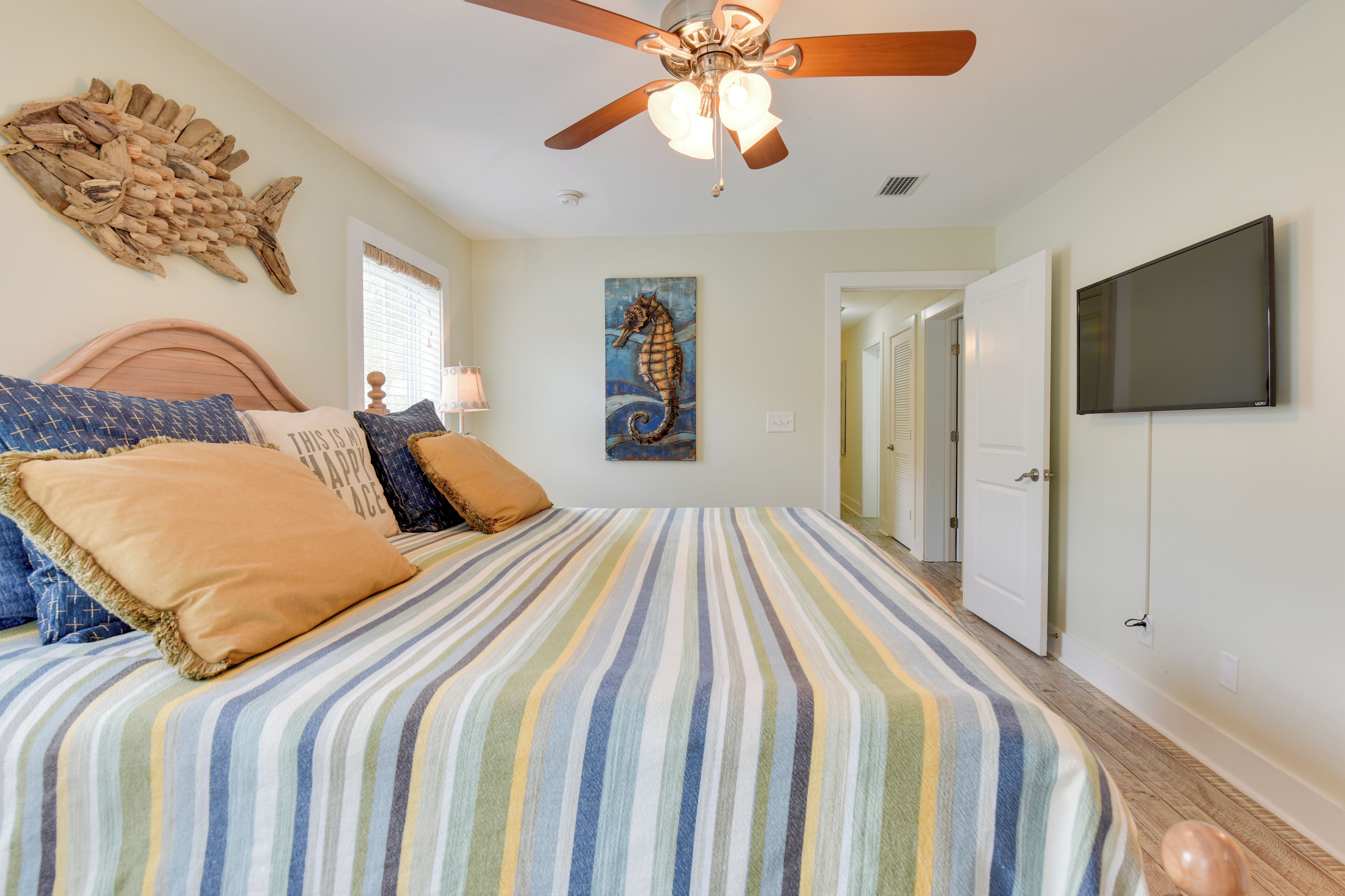 A bedroom with a striped bedspread, a ceiling fan, a mounted TV, and a seahorse painting.