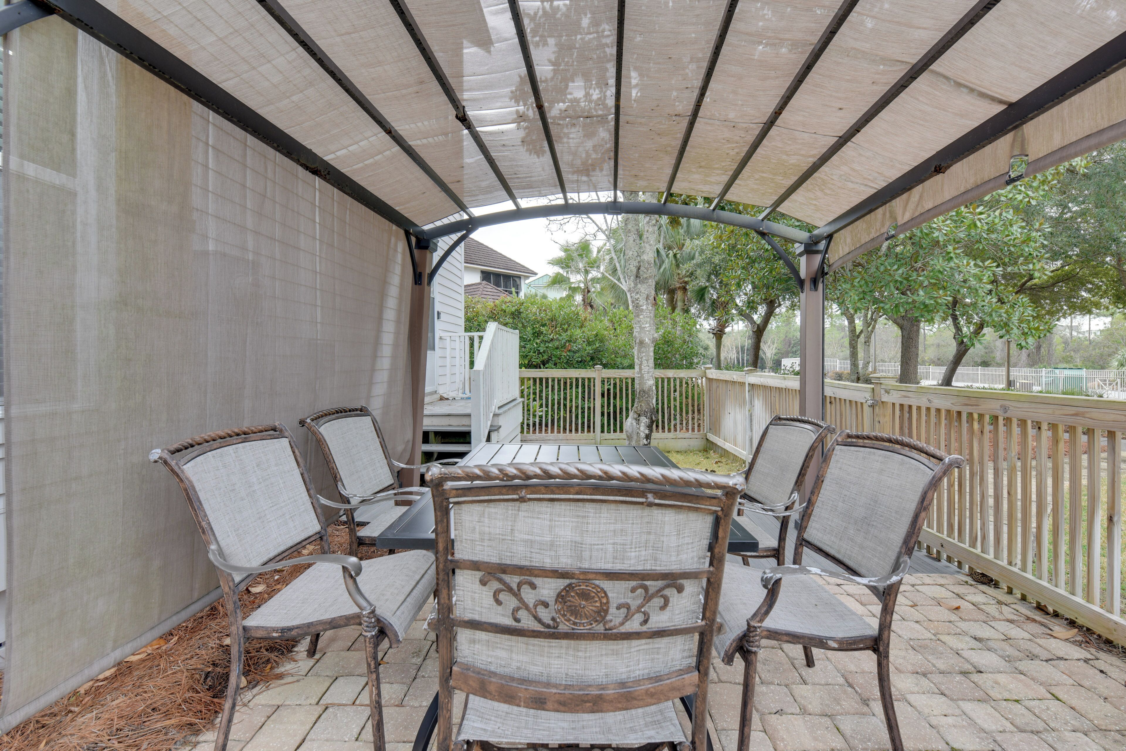 A covered patio with four wicker chairs and a table, surrounded by a wooden fence and trees.