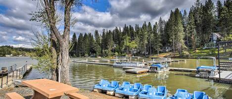 A lakeside area with a row of numbered blue boats, a picnic table, and a dock with a small building in the background.