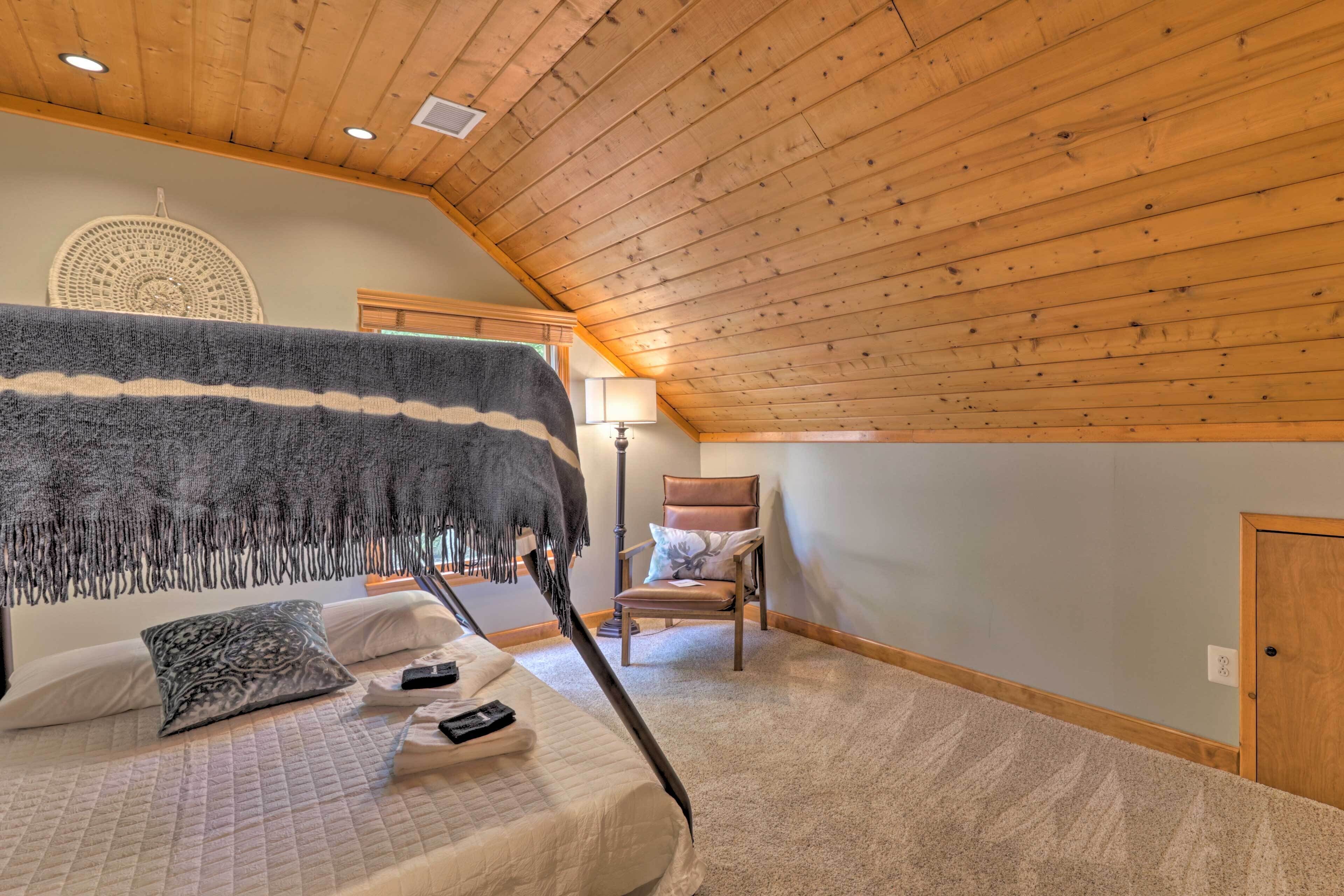 Un dormitorio con techo inclinado, una cama con manta, una silla y una lámpara fijada a la pared.