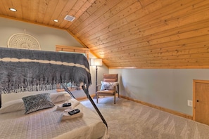 A bedroom with a sloped ceiling, a bed with a blanket, a chair, and a wall-mounted lamp.