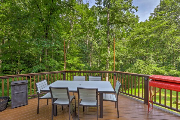 A wooden deck with a table and chairs, surrounded by a railing, overlooking a forested area.