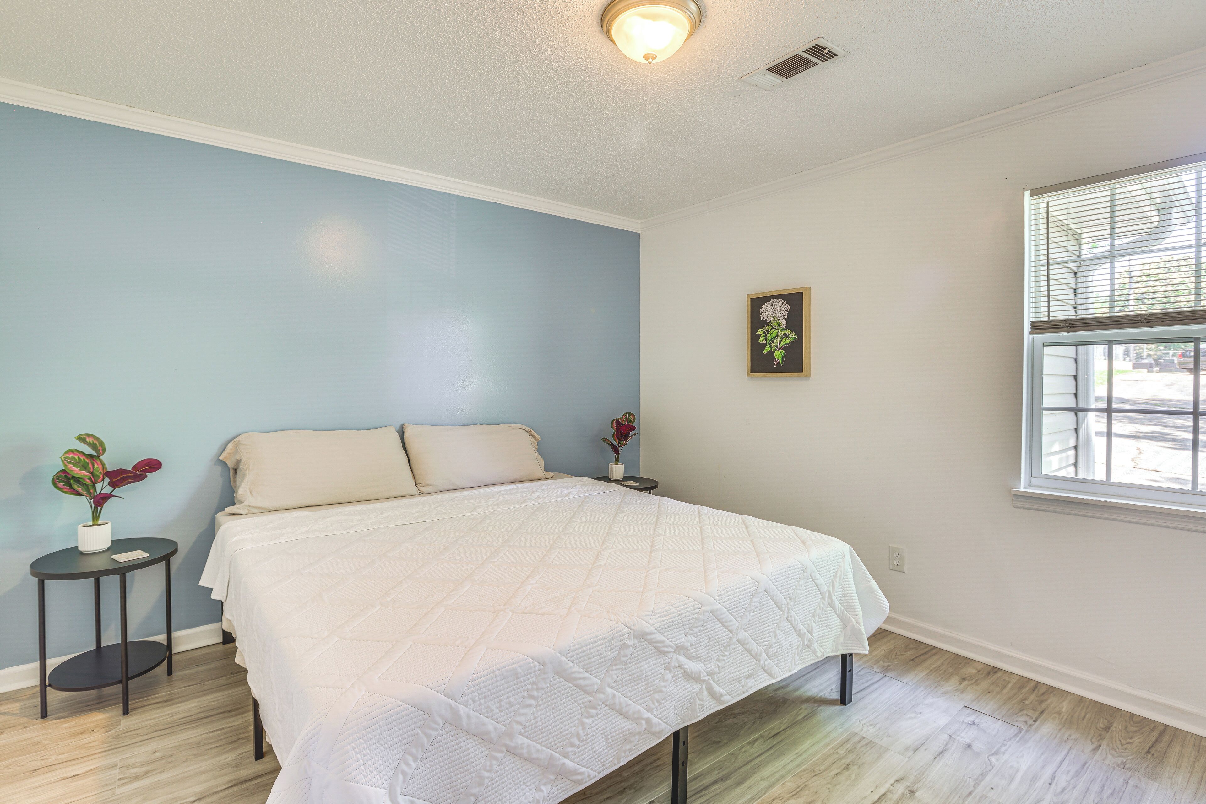 A bedroom with a bed, a small table with a plant, a picture on the wall, and a window.