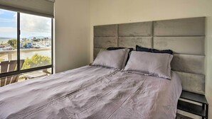 Quarto com cama grande, cabeceira cinza e vista para uma marina através de uma janela.