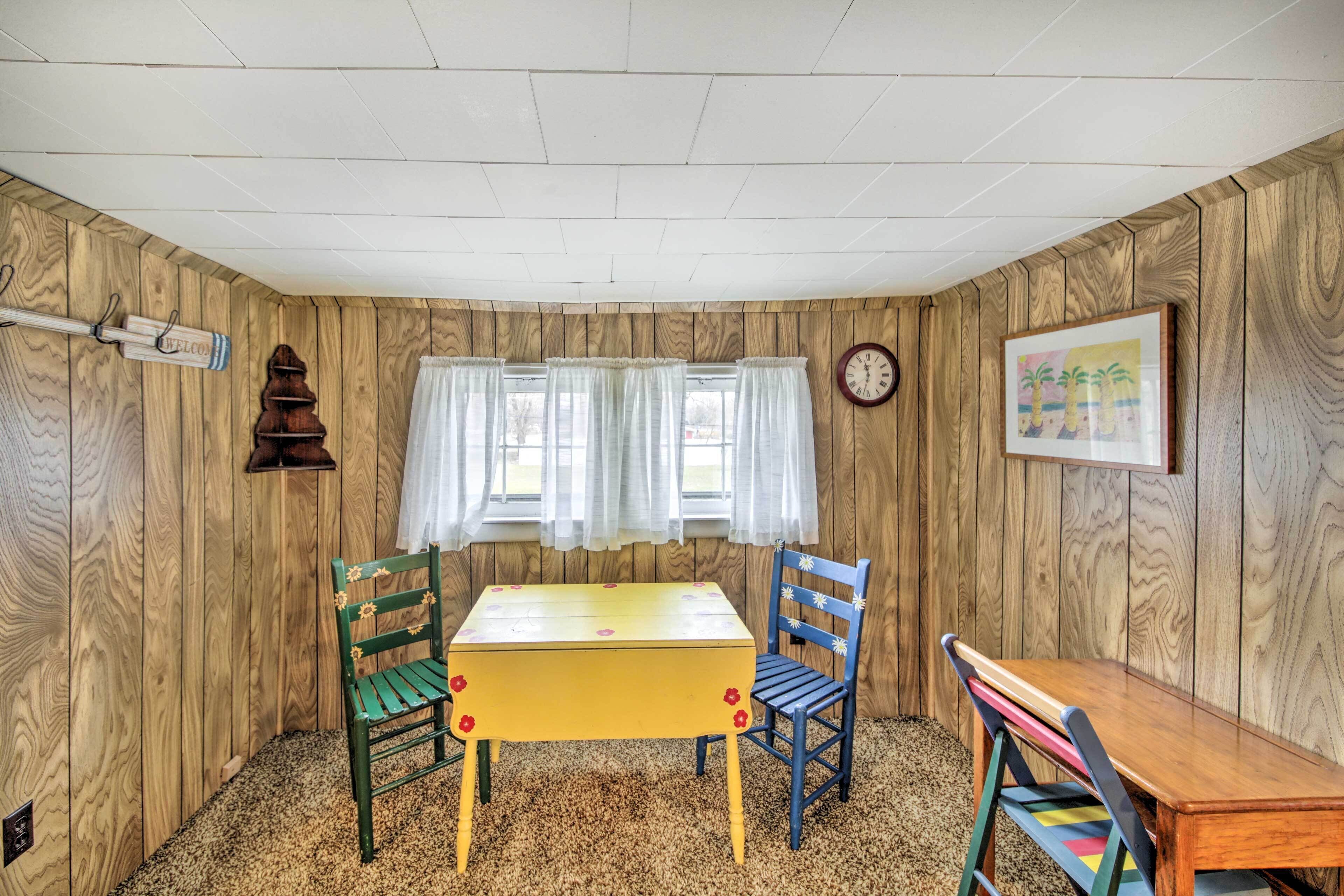A room with wooden walls, a yellow table, green and blue chairs, a clock, and a window with curtains.