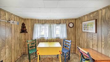 A room with wooden walls, a yellow table, green and blue chairs, a clock, and a window with curtains.