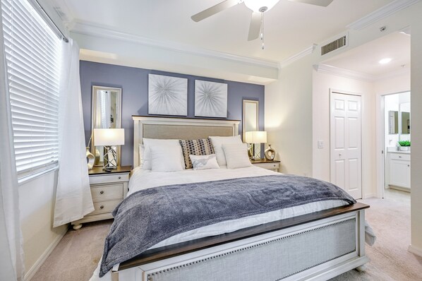 A bedroom with a large bed, two bedside tables with lamps, a ceiling fan, and a door leading to another room.