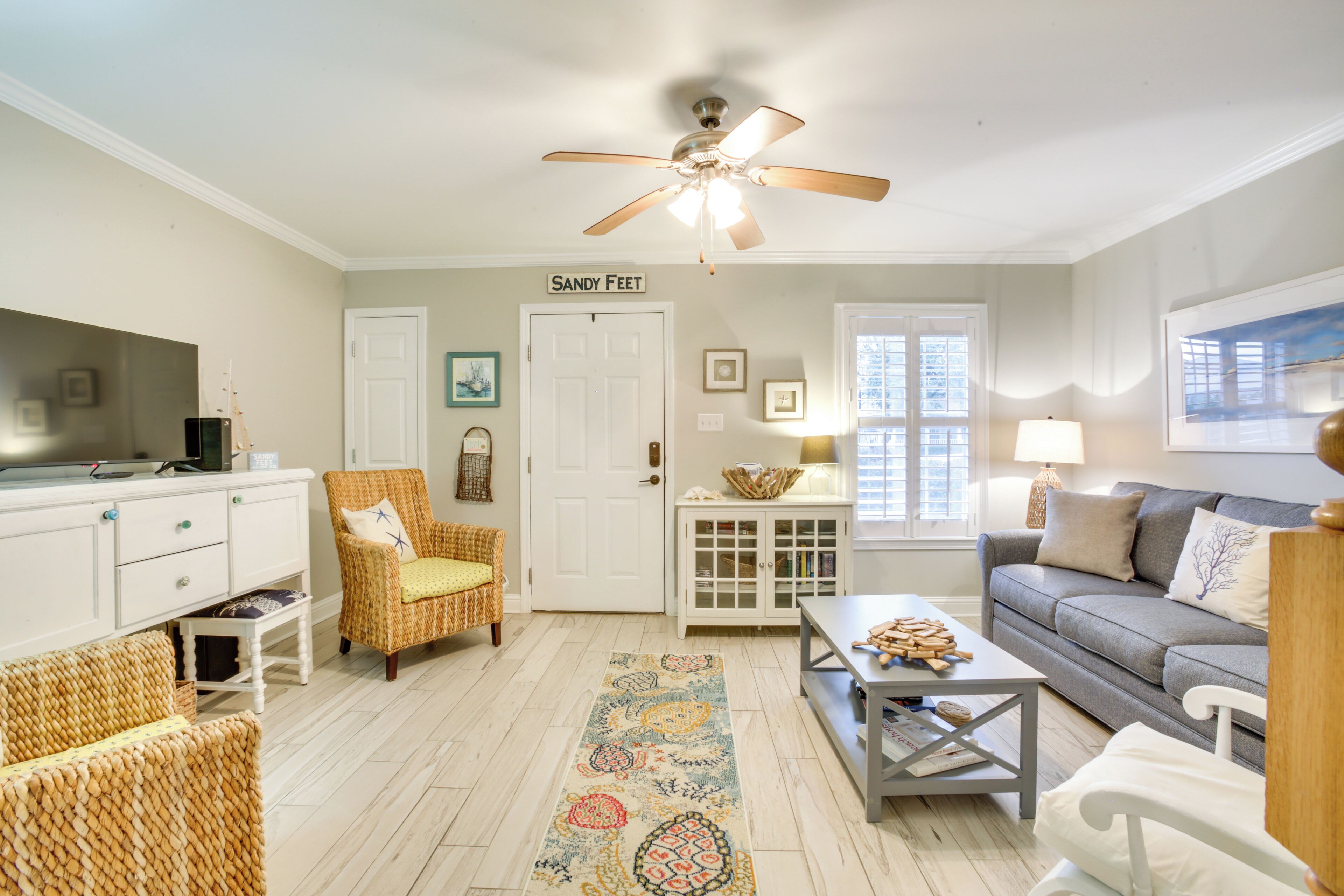 A living room with a ceiling fan, a sofa, a coffee table, wicker chairs, a television, and a door labeled "Sandy Feet".