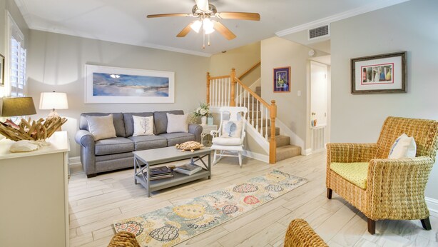 A living room with a gray sofa, a coffee table, a wicker armchair, a ceiling fan, and a staircase. - Old Town Bay St Louis Townhome: Walk to Beach (Bay St Louis)