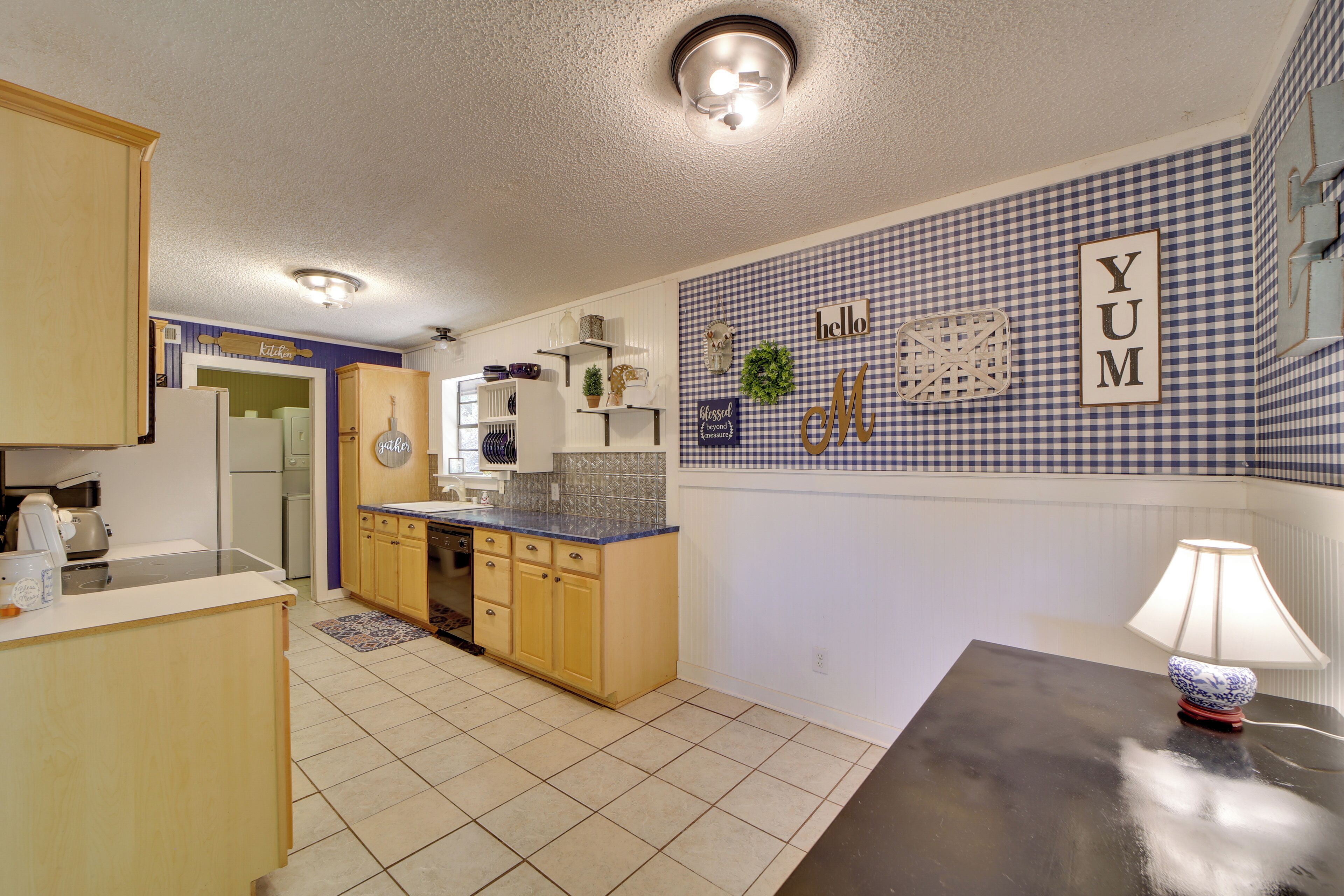 A kitchen with light wooden cabinets, a black countertop, and a tiled backsplash. There is a wall-mounted shelf with decorative items and a wall clock.