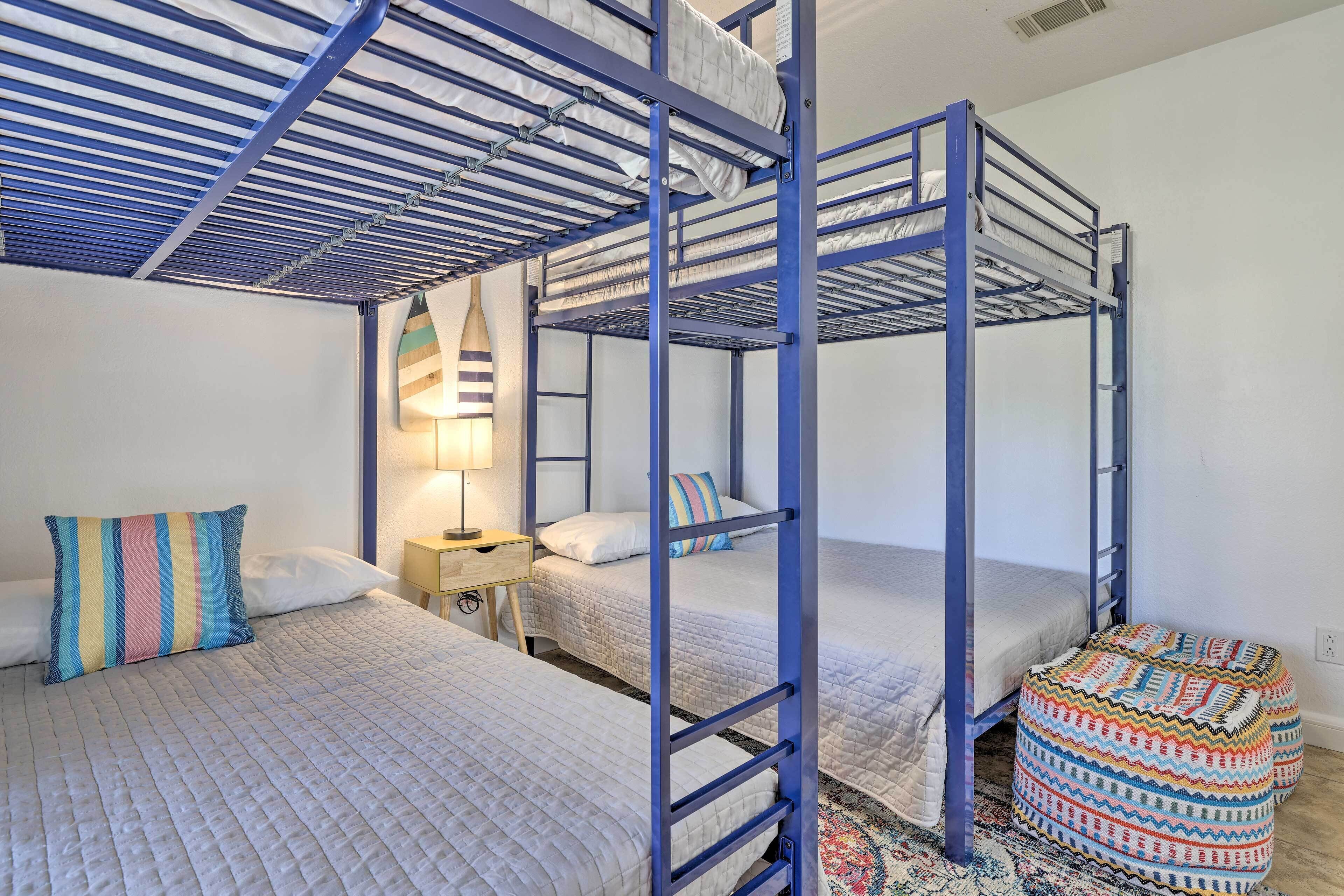 A bunk bed room with a blue metal frame, a bedside table with a lamp, and a colorful ottoman.