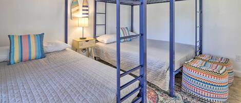 A bunk bed room with a blue metal frame, a bedside table with a lamp, and a colorful ottoman.