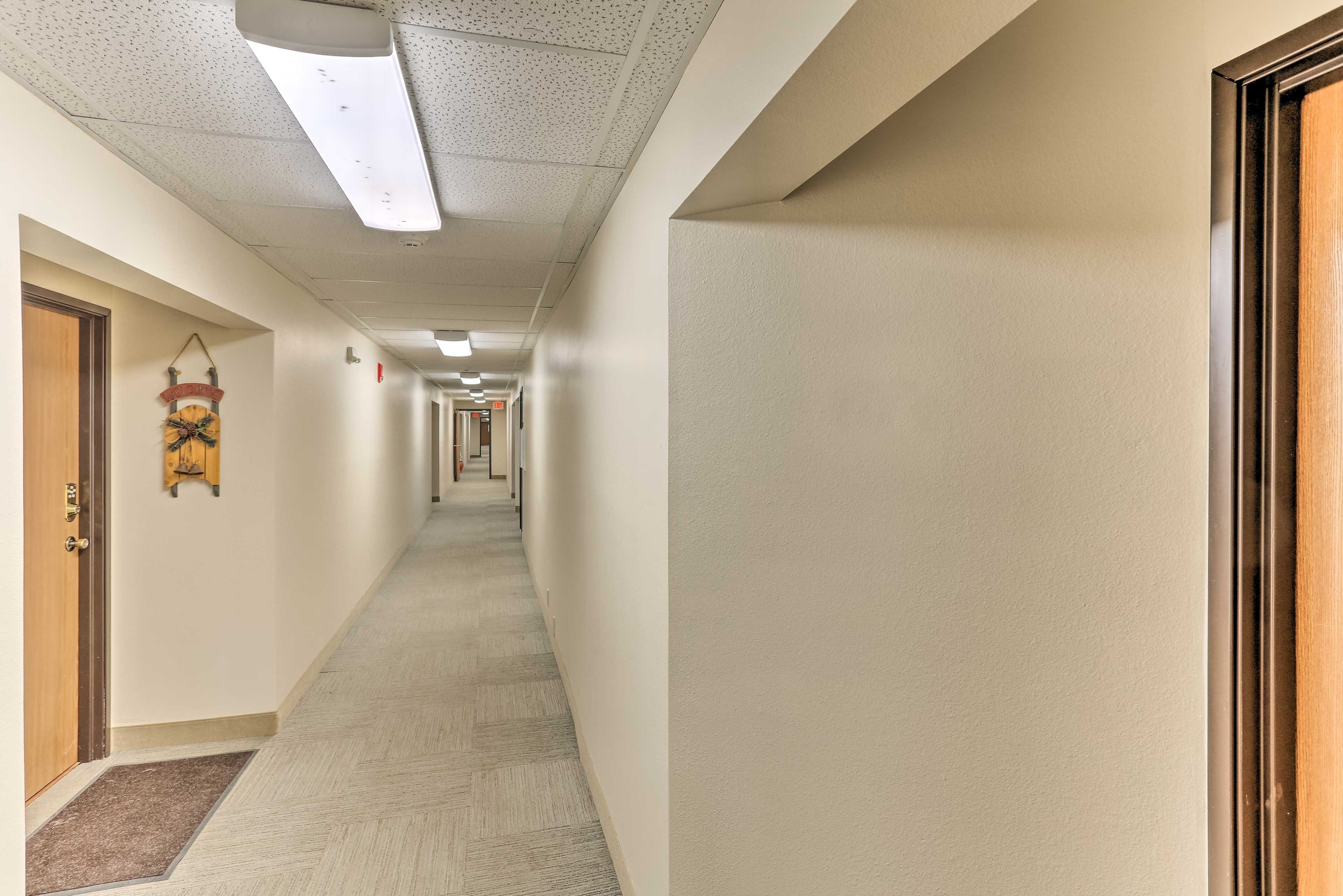 A long, narrow hallway with a carpeted floor, white walls, and a ceiling with fluorescent lighting.