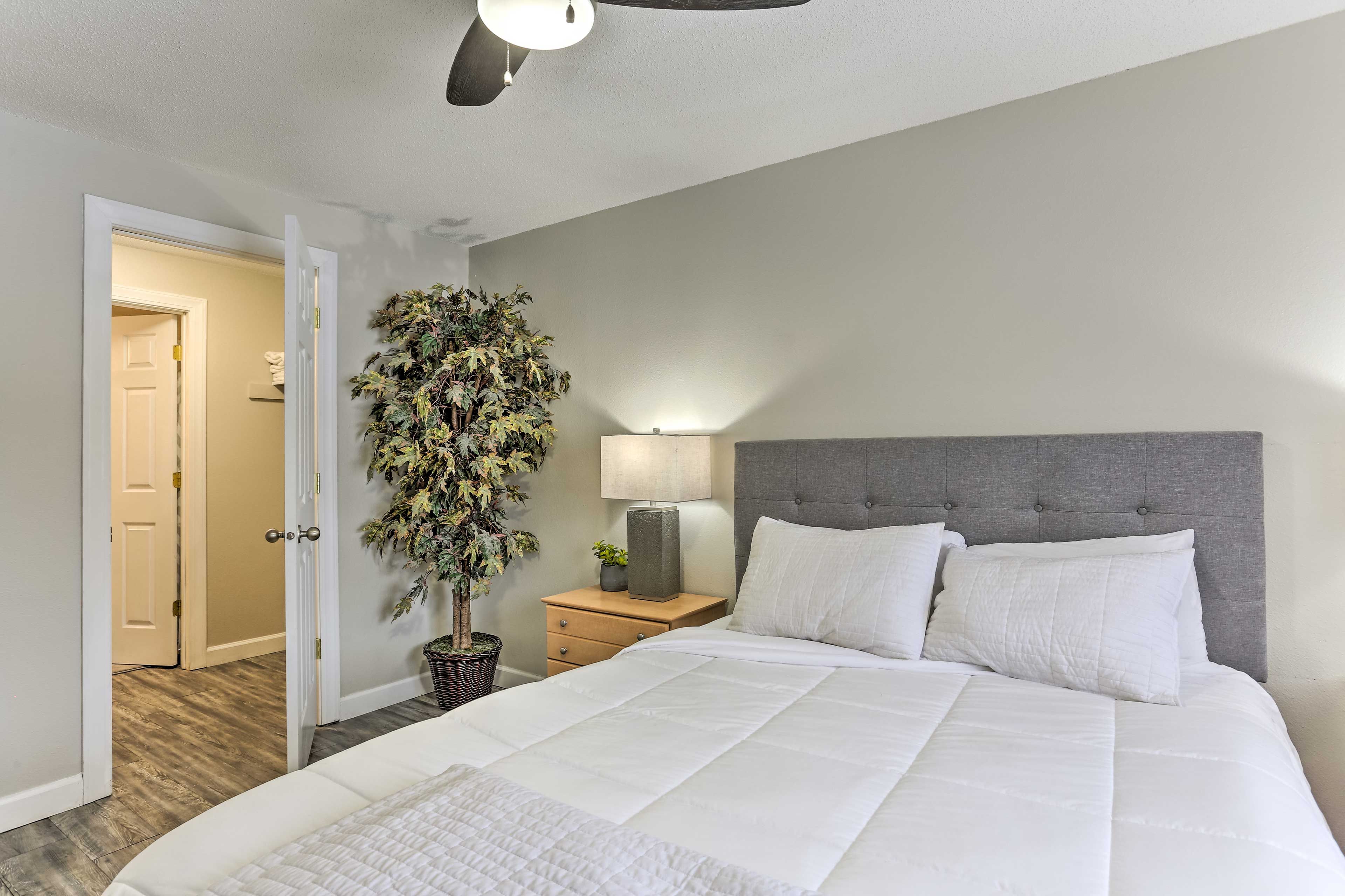 A bedroom with a large bed, a nightstand with a lamp, a potted plant, and a door leading to another room.
