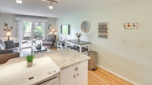 Una cocina moderna con encimera de granito, armarios blancos y una estufa integrada. Hay una zona de comedor con mesa y sillas, y una sala de estar con sofá y mesa de centro.